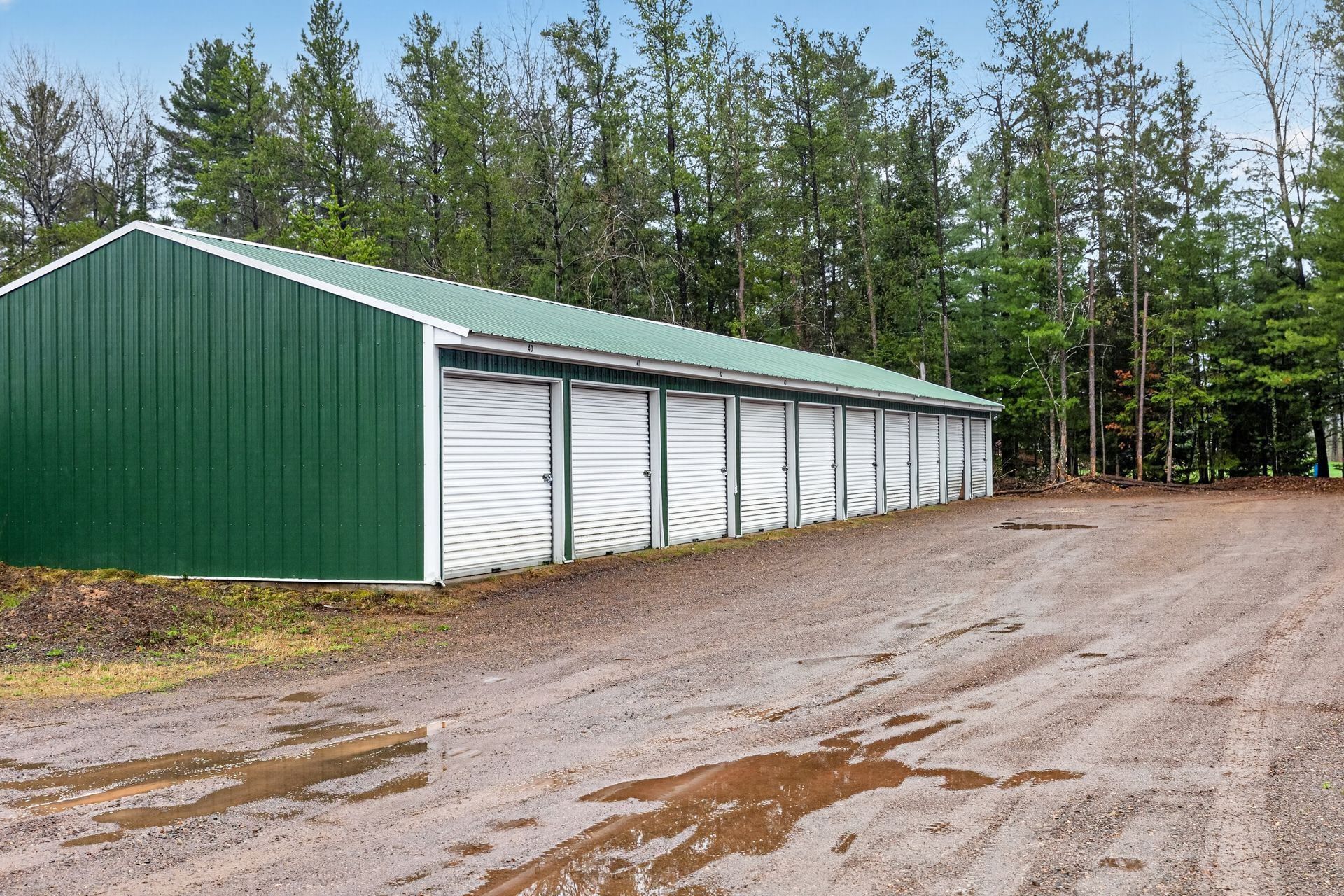 Green metal storage building with white roll-up doors on a gravel lot by trees