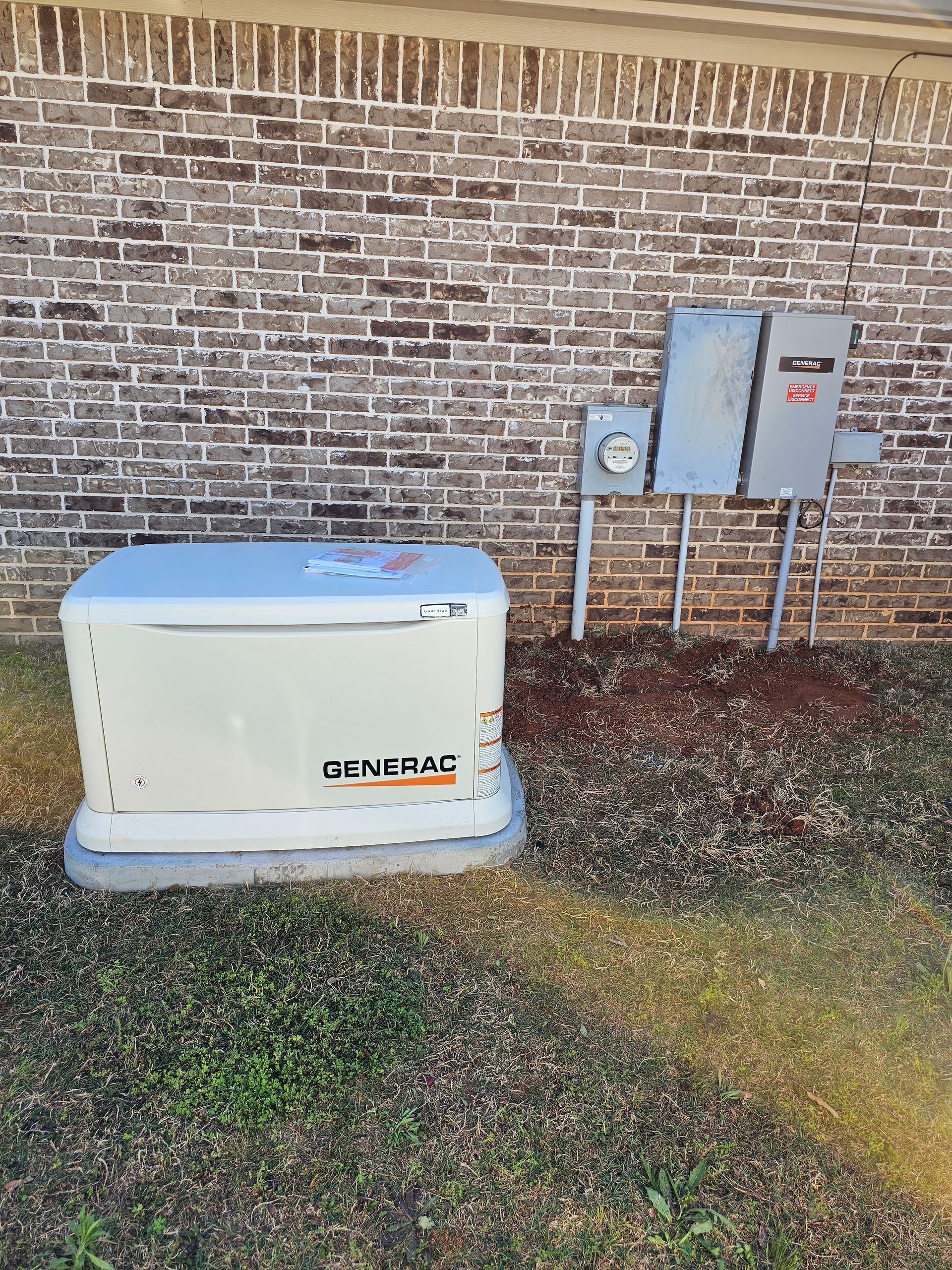 A generator is sitting in the grass next to a brick wall.