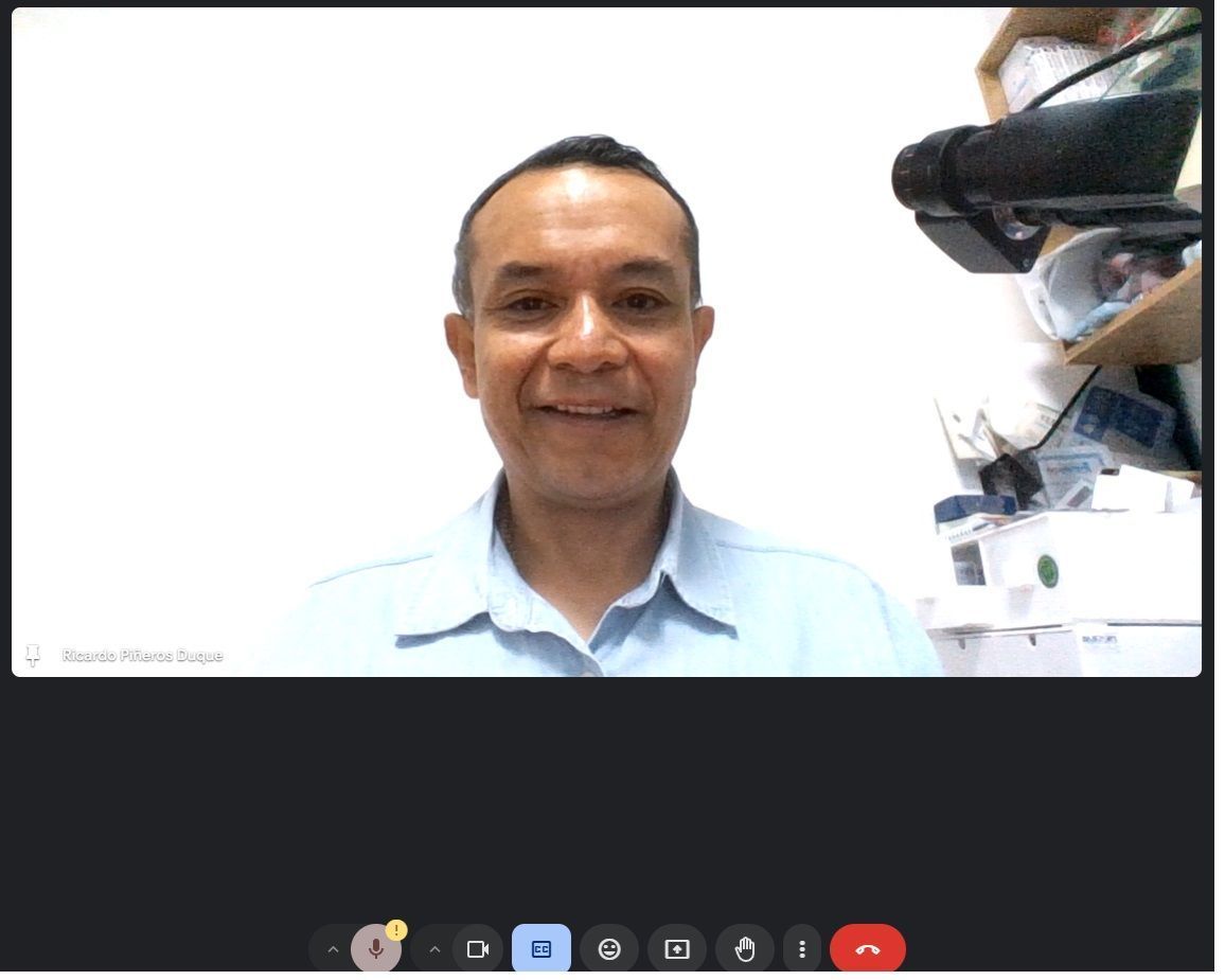 Hombre sonriendo en una videollamada; fondo blanco, camisa azul suave y un micrófono.