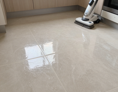 A robot vacuum cleans a shiny beige tiled floor near a wall and window reflection.