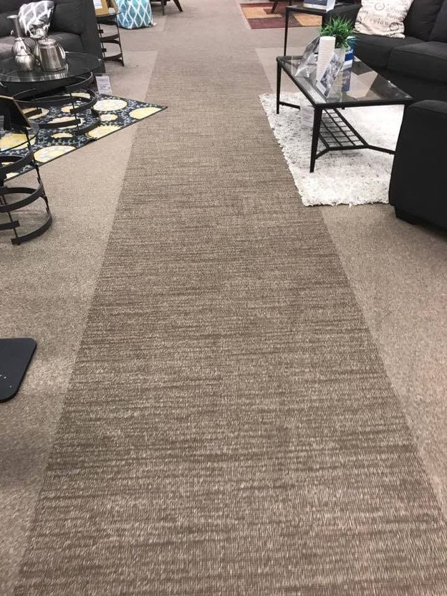 Carpeted aisle in a furniture store, with rugs and tables displayed on both sides.
