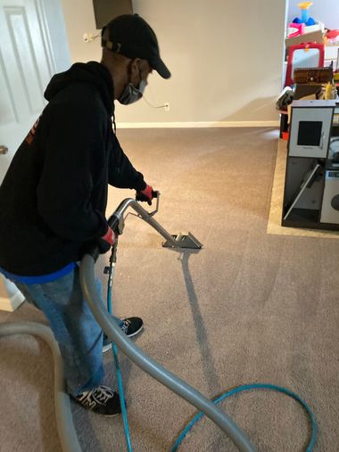 Person vacuuming carpet in a room, wearing dark clothes and a cap.