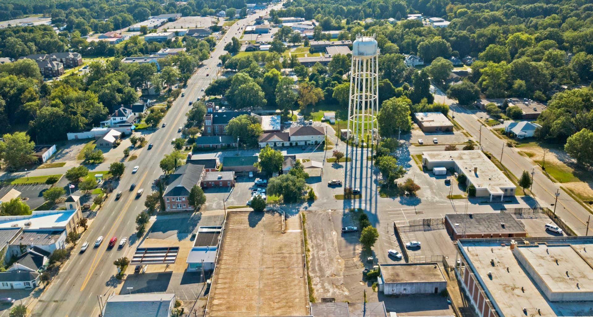 Aerial view of a town with a tall water tower. Buildings and roads are visible, surrounded by green trees.
