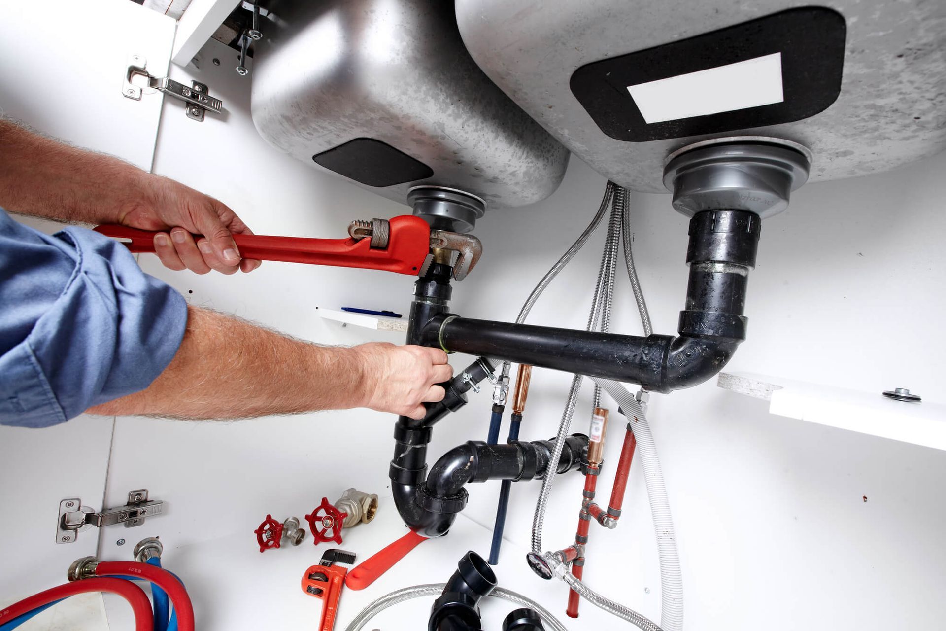 Plumber using a wrench under a kitchen sink, fixing pipes in a white cabinet.