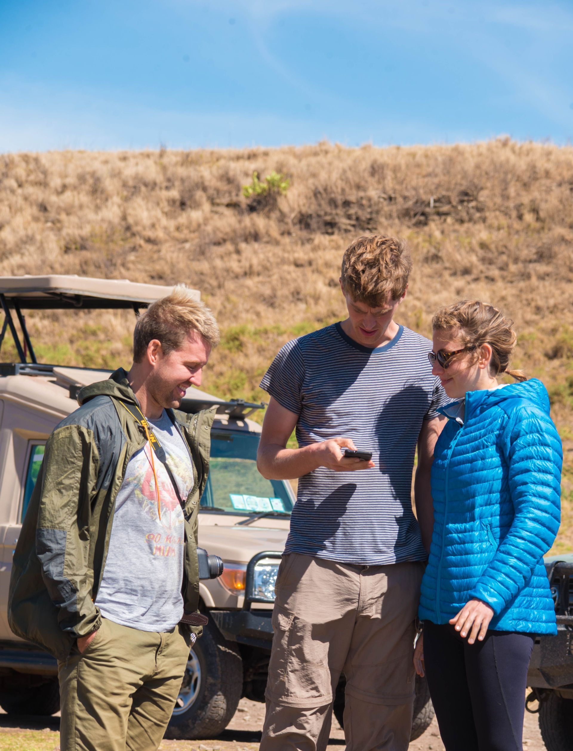 Three people standing outdoors by a vehicle, looking at a phone under a blue sky.