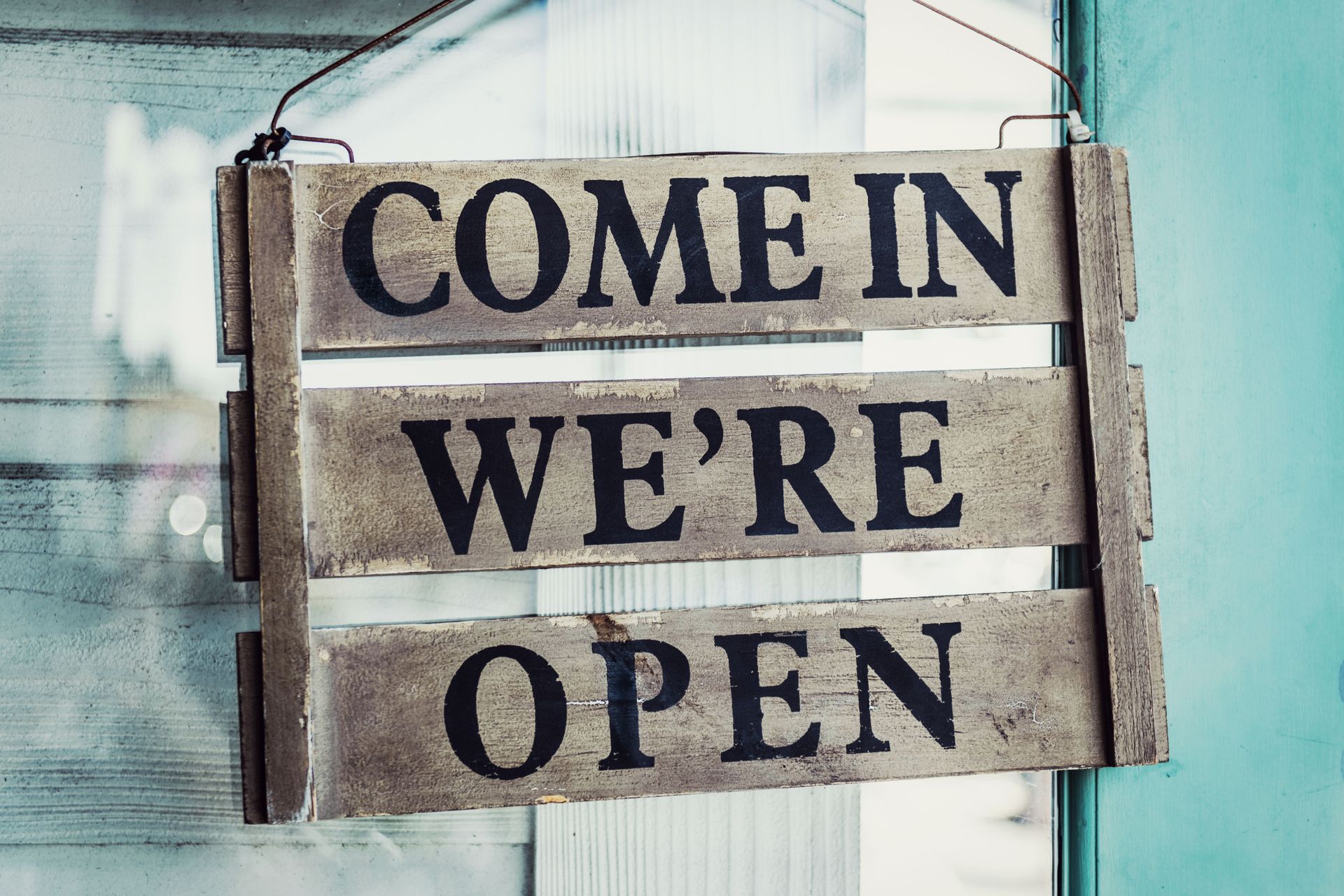 Wooden sign hanging in a window reads “COME IN WE’RE OPEN”