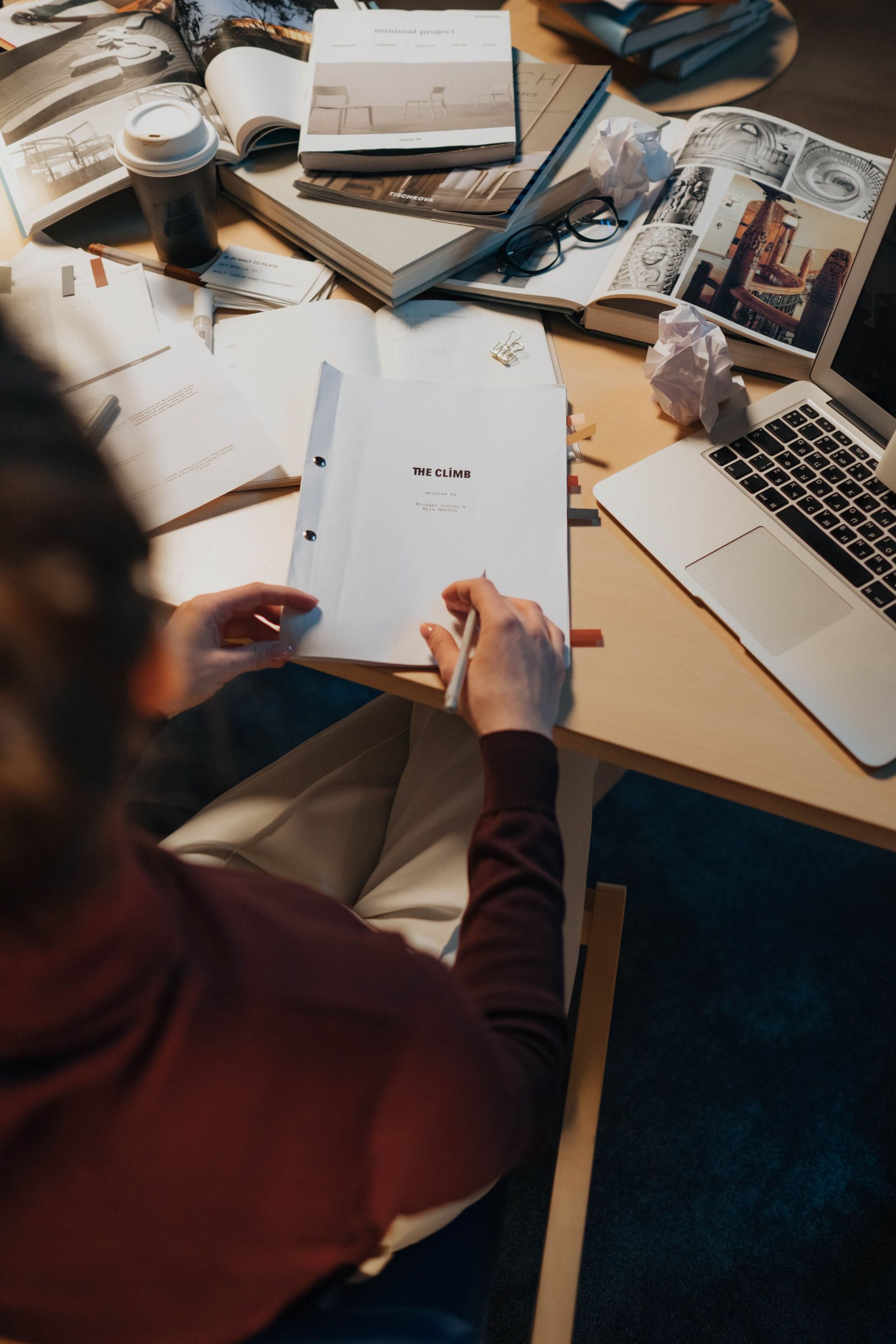 Person holding a booklet at a cluttered desk with laptop, notebooks, and magazines