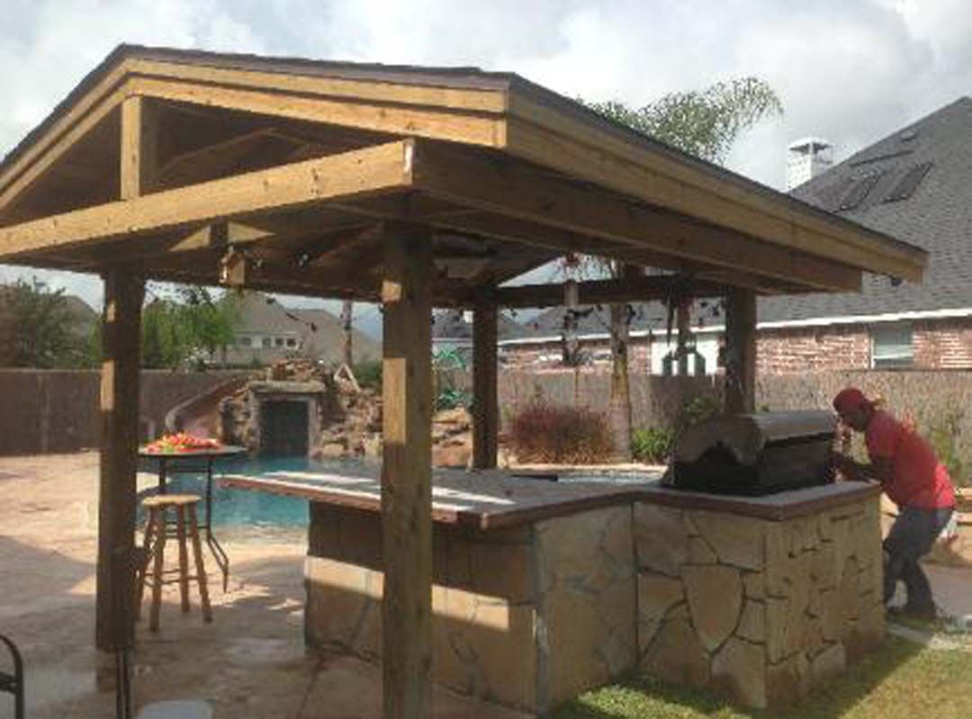 A man is working on a grill under a wooden gazebo next to a pool.