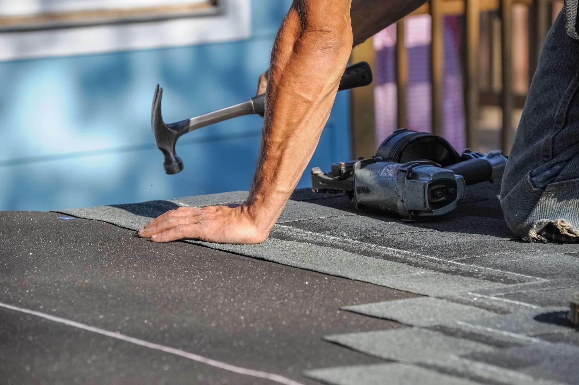 Worker nailing roofing shingles with a hammer on a rooftop, with a power tool nearby