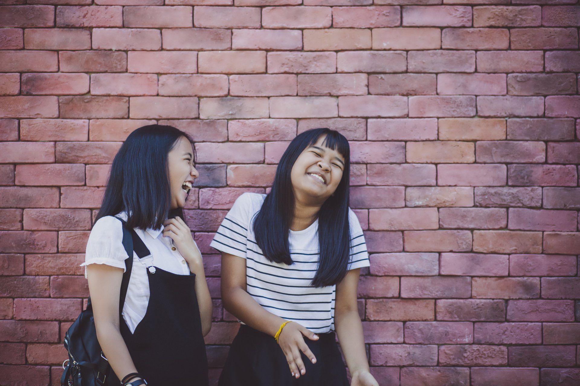 Two people laughing in front of a brick wall. One person wears overalls, the other a striped shirt.