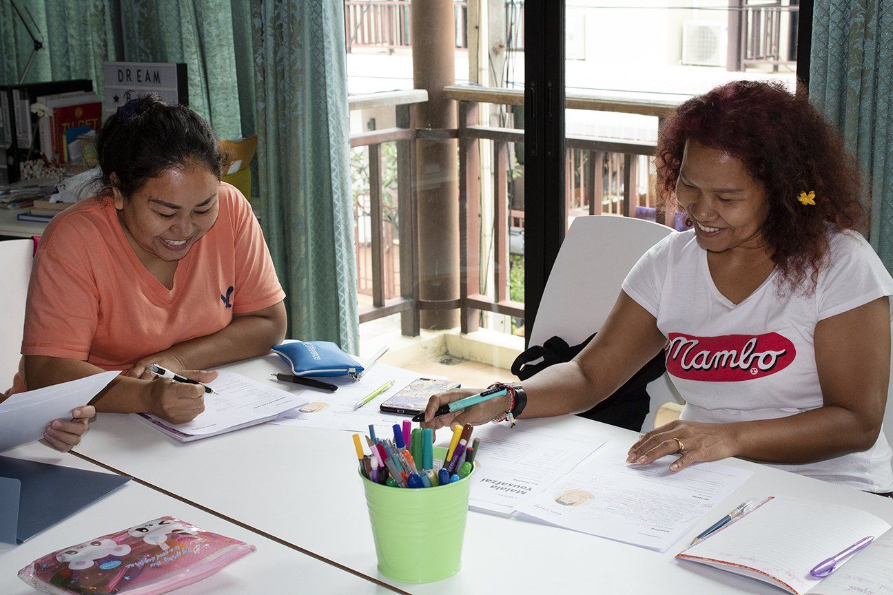 Two women sit at a table, smiling and working on papers. Colorful pens in a green cup. Balcony in background.
