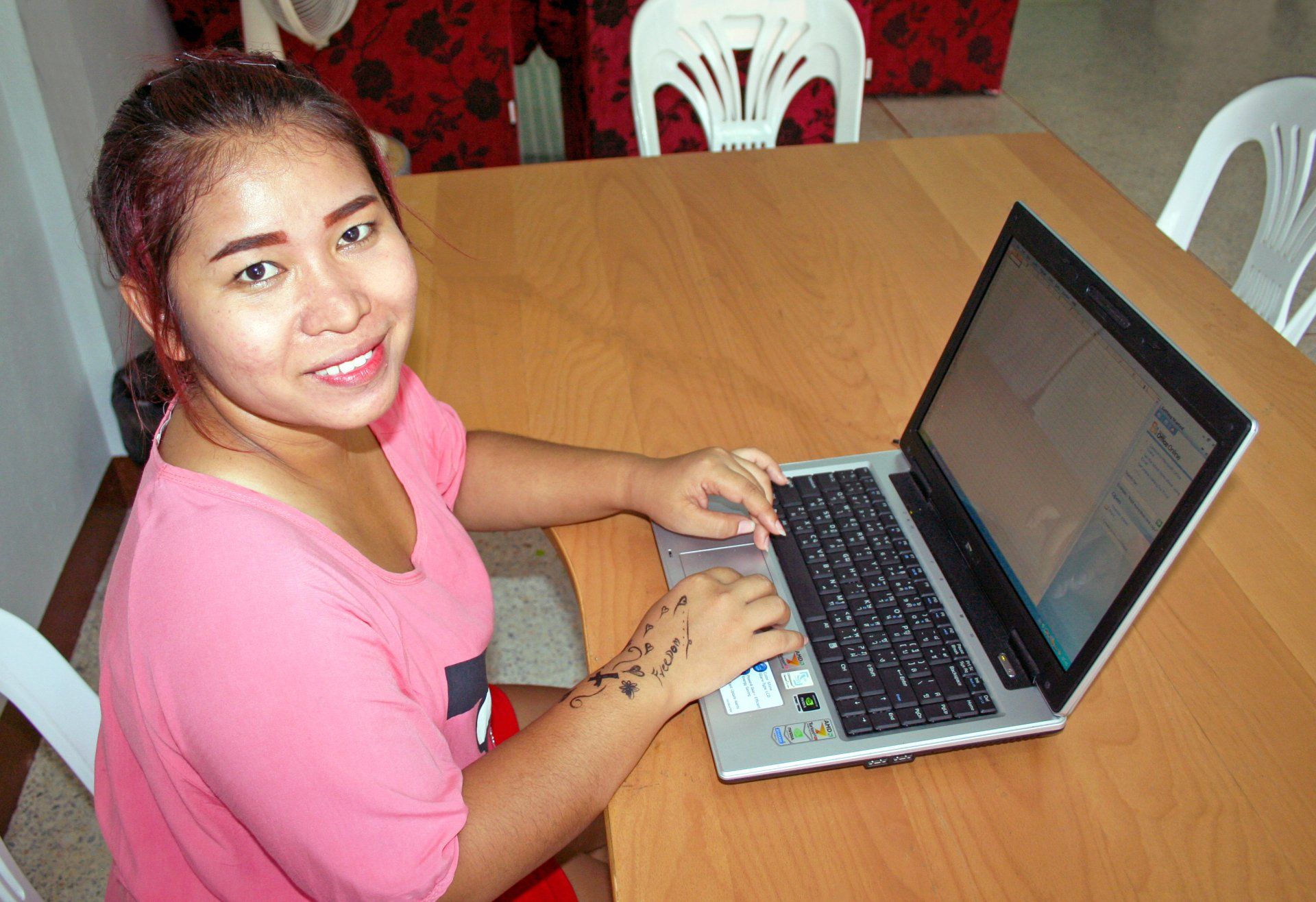 Woman in pink shirt smiles while using a laptop at a wooden table.
