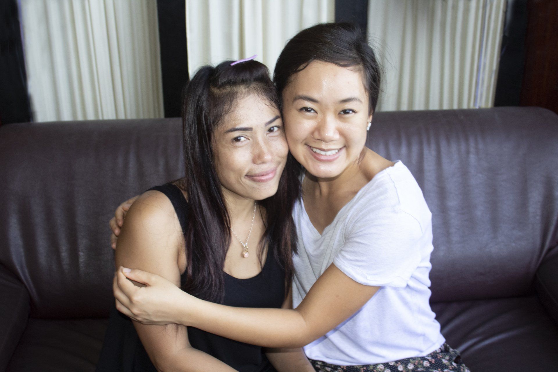 Two people smiling and hugging on a brown couch. One wears black, the other, white.