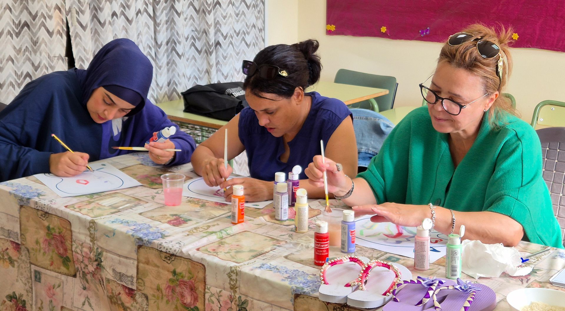 Three people painting at a table. Two women are focused on their art, while the other looks on.