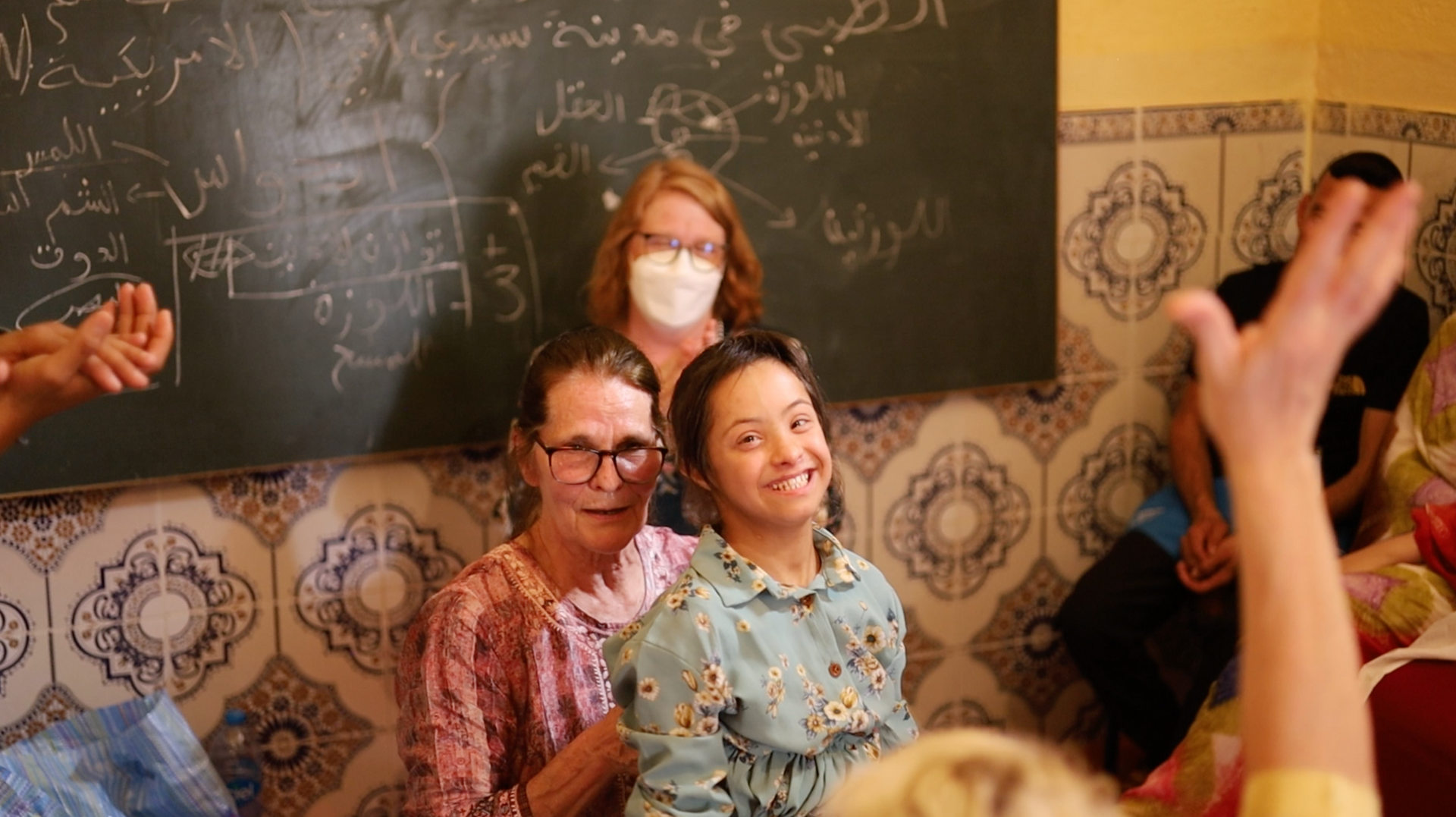 Smiling woman in blue floral shirt with another woman and a person with an arm raised near a chalkboard.