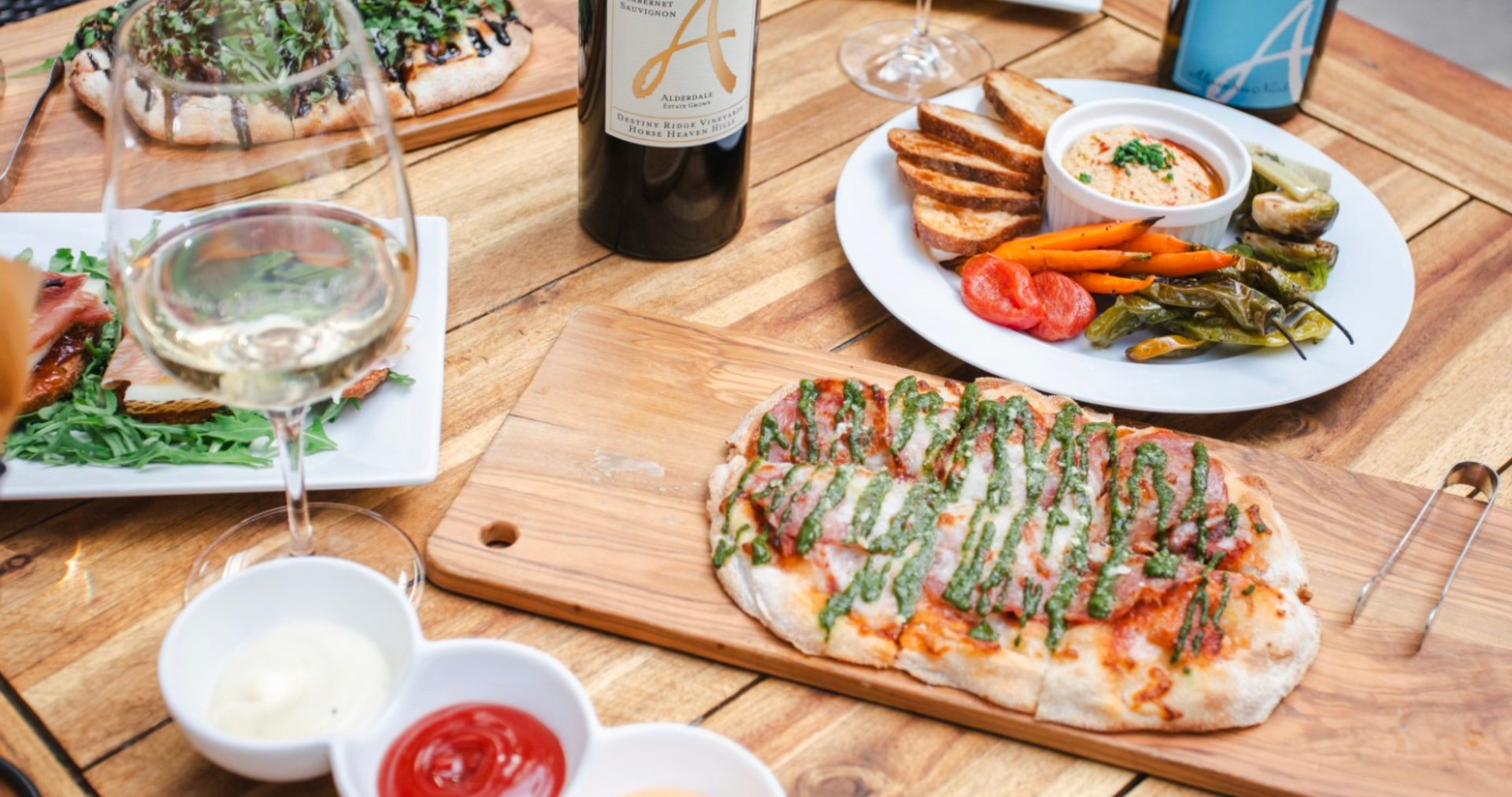 Gourmet food spread with flatbread, vegetables, dips, wine bottles, and glasses on a wooden table.