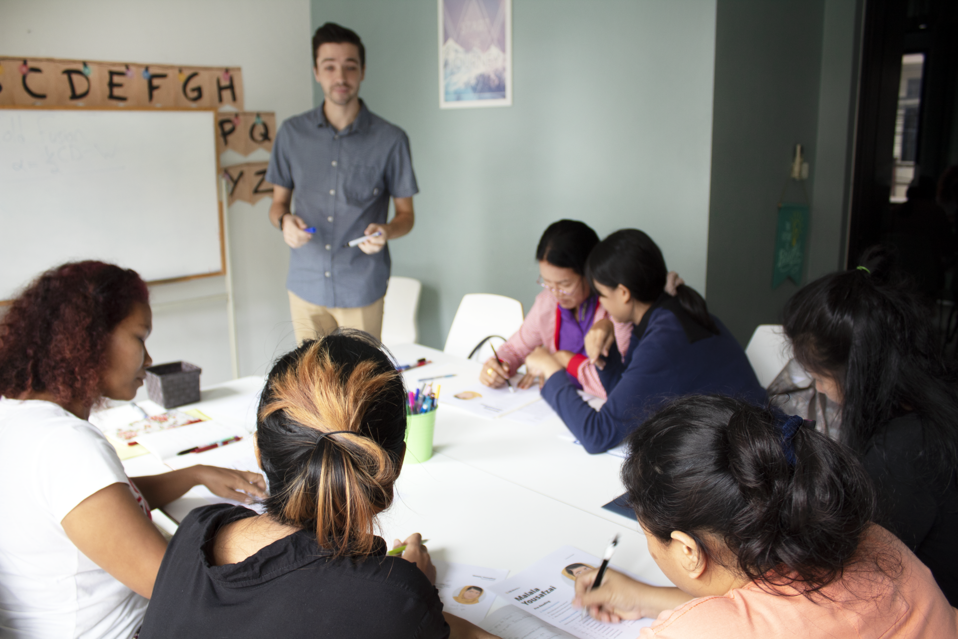 Teacher leading a class of students at a table. Students are taking notes, white board with letters on the wall.