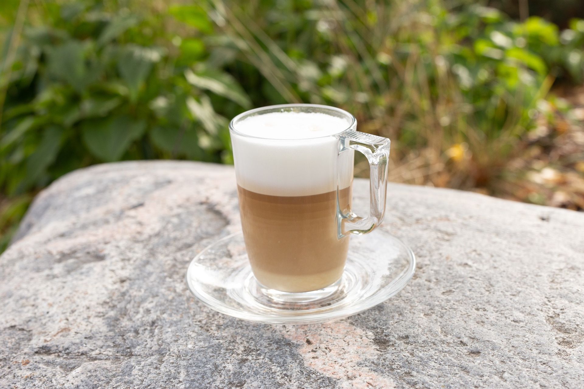 Ant akmens lauke stovi stiklinė latte su puta ant lėkštutės.