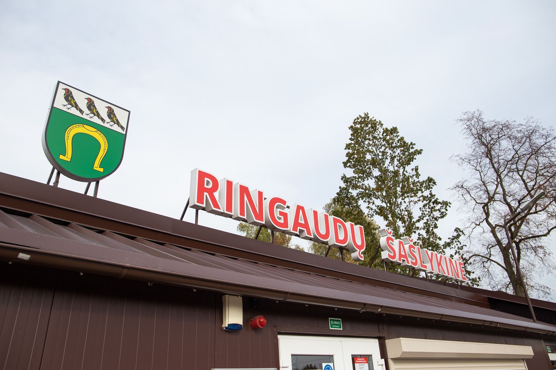 RINGAUDŲ ženklas ant rudo pastato su žaliai baltu herbu, už jo medžiai