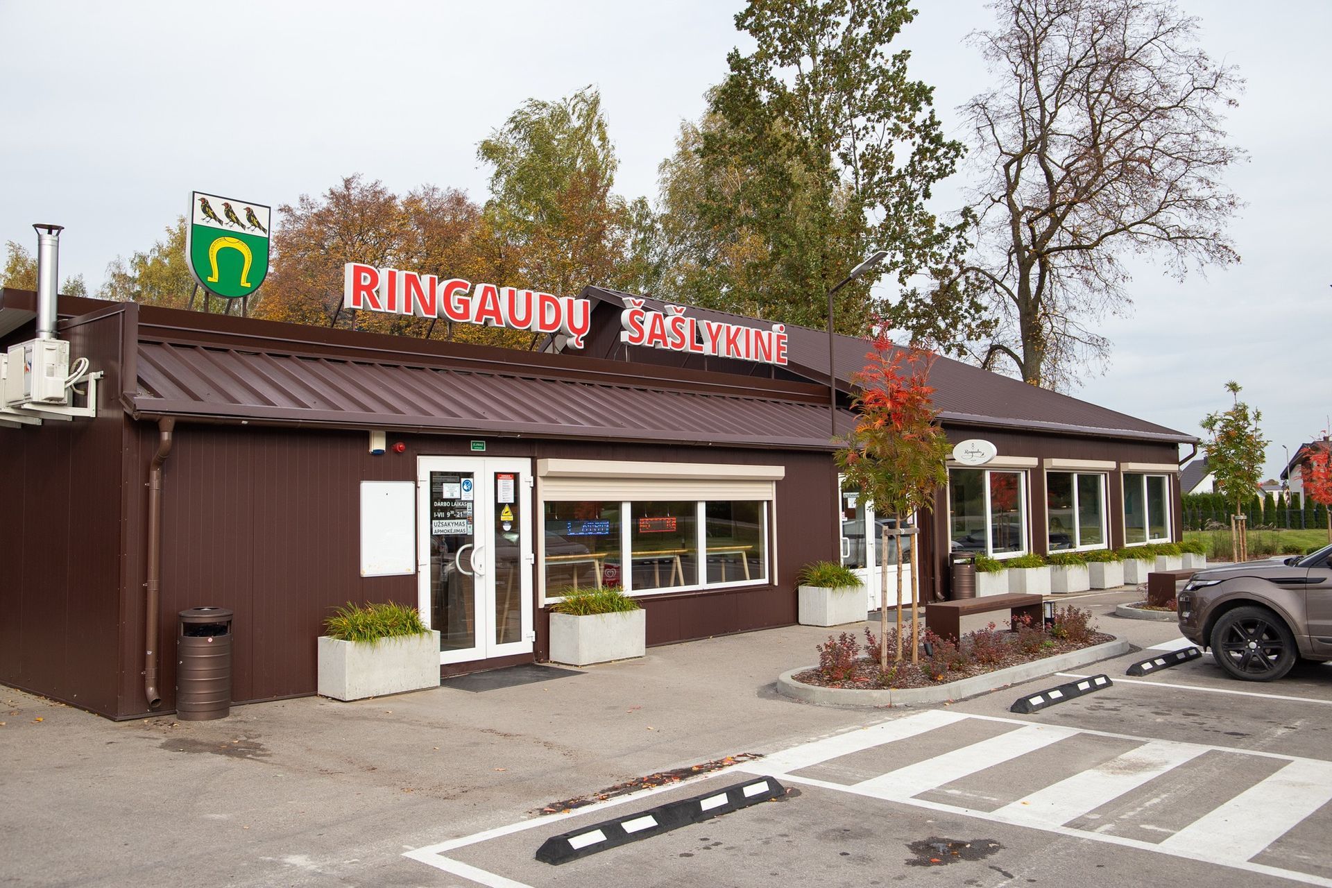 Rudas pastatas su raudonu ženklu ir baltais langais šalia pėsčiųjų perėjos ir stovinčio sunkvežimio