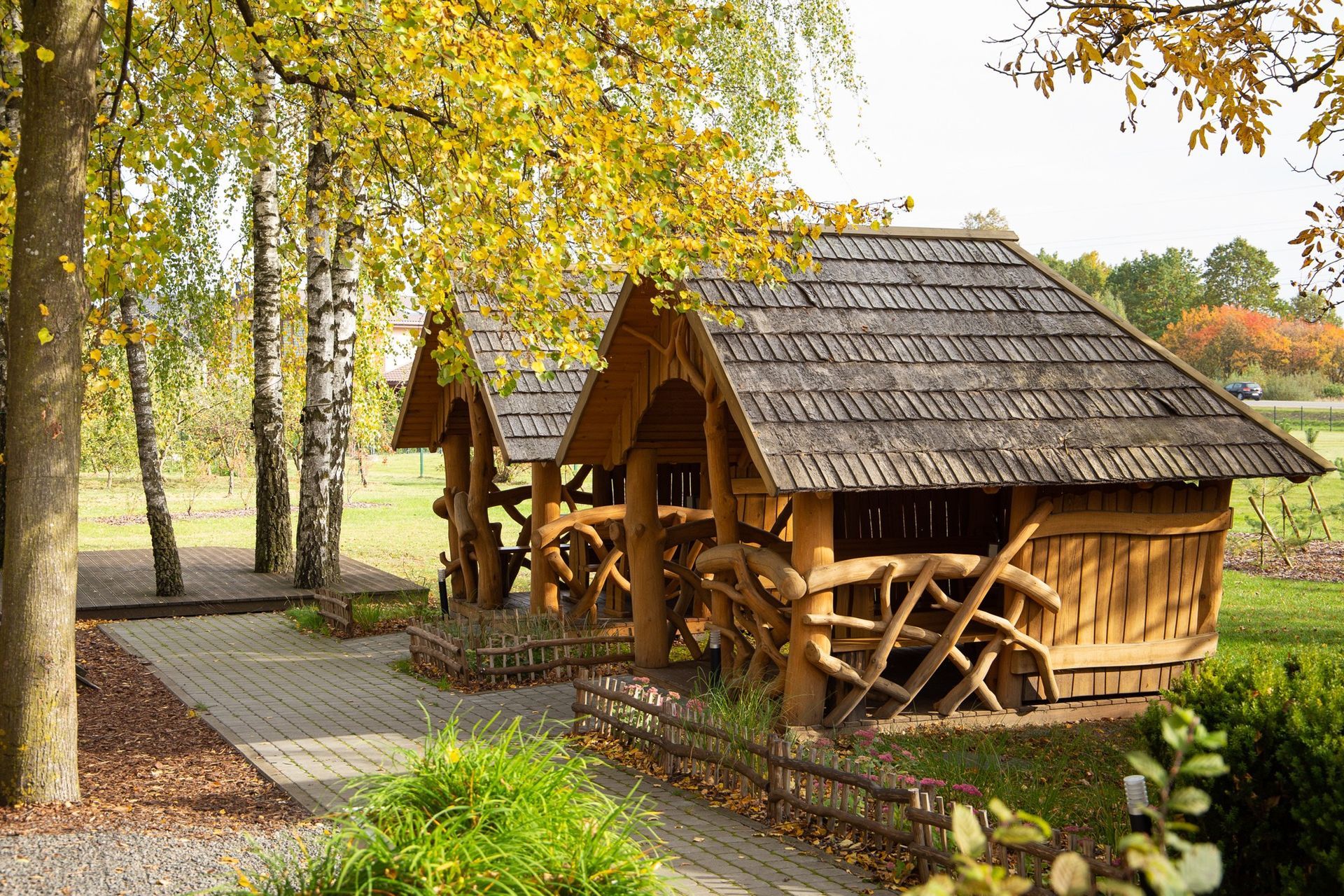 Mediniai kaimiški nameliai saulėtame rudens miške su akmeniniu taku ir medžiais.