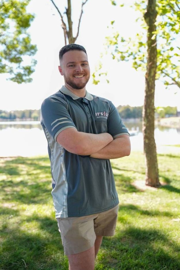Man standing with arms crossed in a gray polo and tan shorts in a sunny park by the water