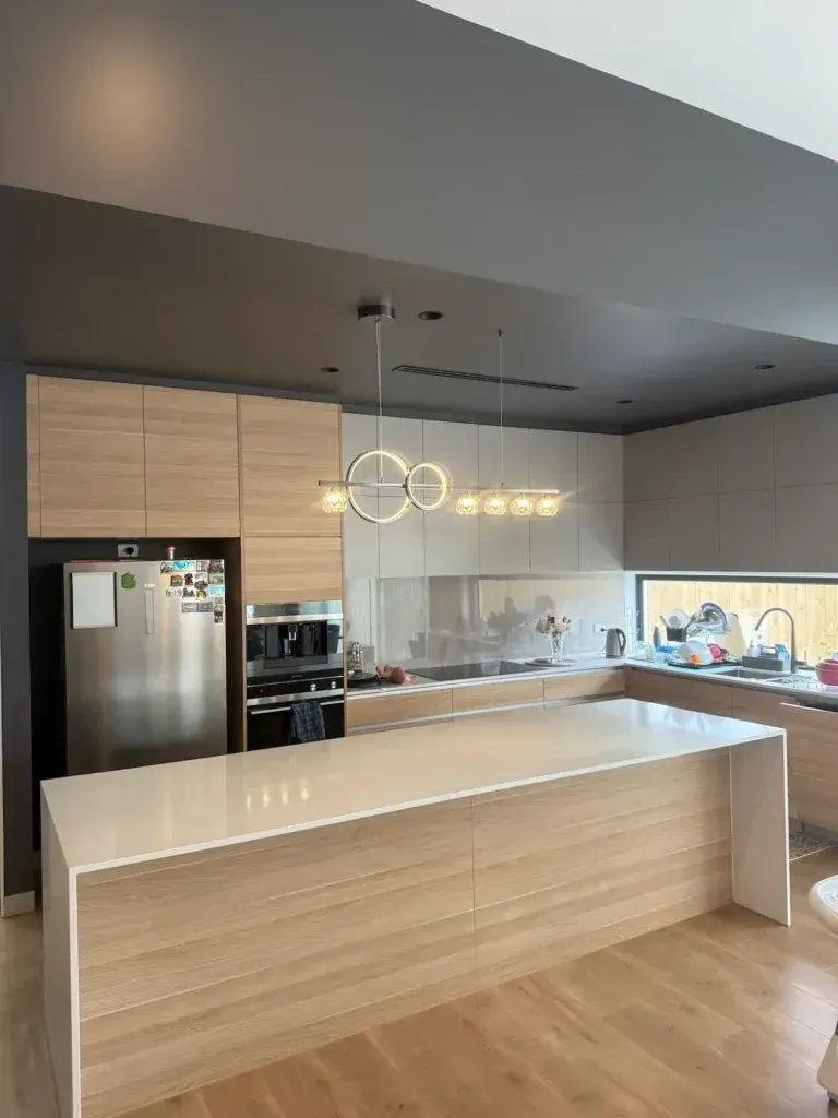 Modern kitchen with light wood cabinets, white countertop, stainless steel appliances, and overhead lighting.