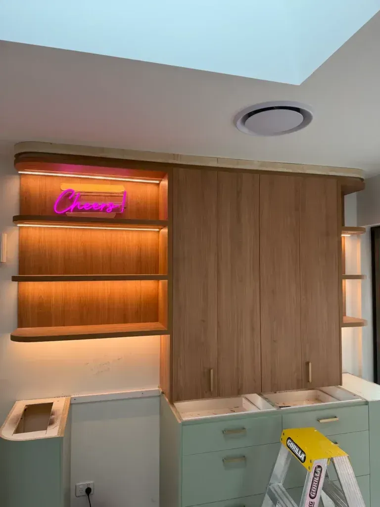 Built-in kitchen shelving and cabinets with warm lighting. A pink neon sign says 