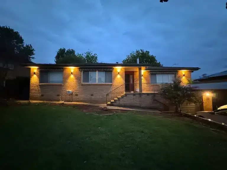 A house at dusk, lit up by warm exterior lights. Green grass and a cloudy sky.