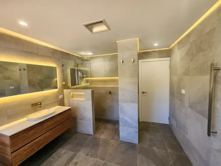 Modern bathroom with gray tile, wood vanity, backlit mirror, and shower area.