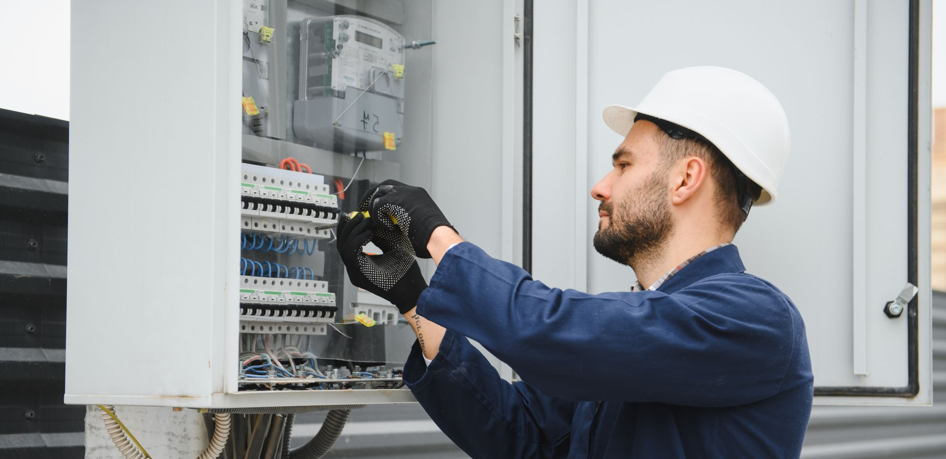 An electrician in a hard hat and work uniform performs maintenance on an open electrical circuit breaker panel.