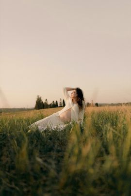 Woman in a white dress reclining in tall grass at sunset, with a soft, hazy sky behind her