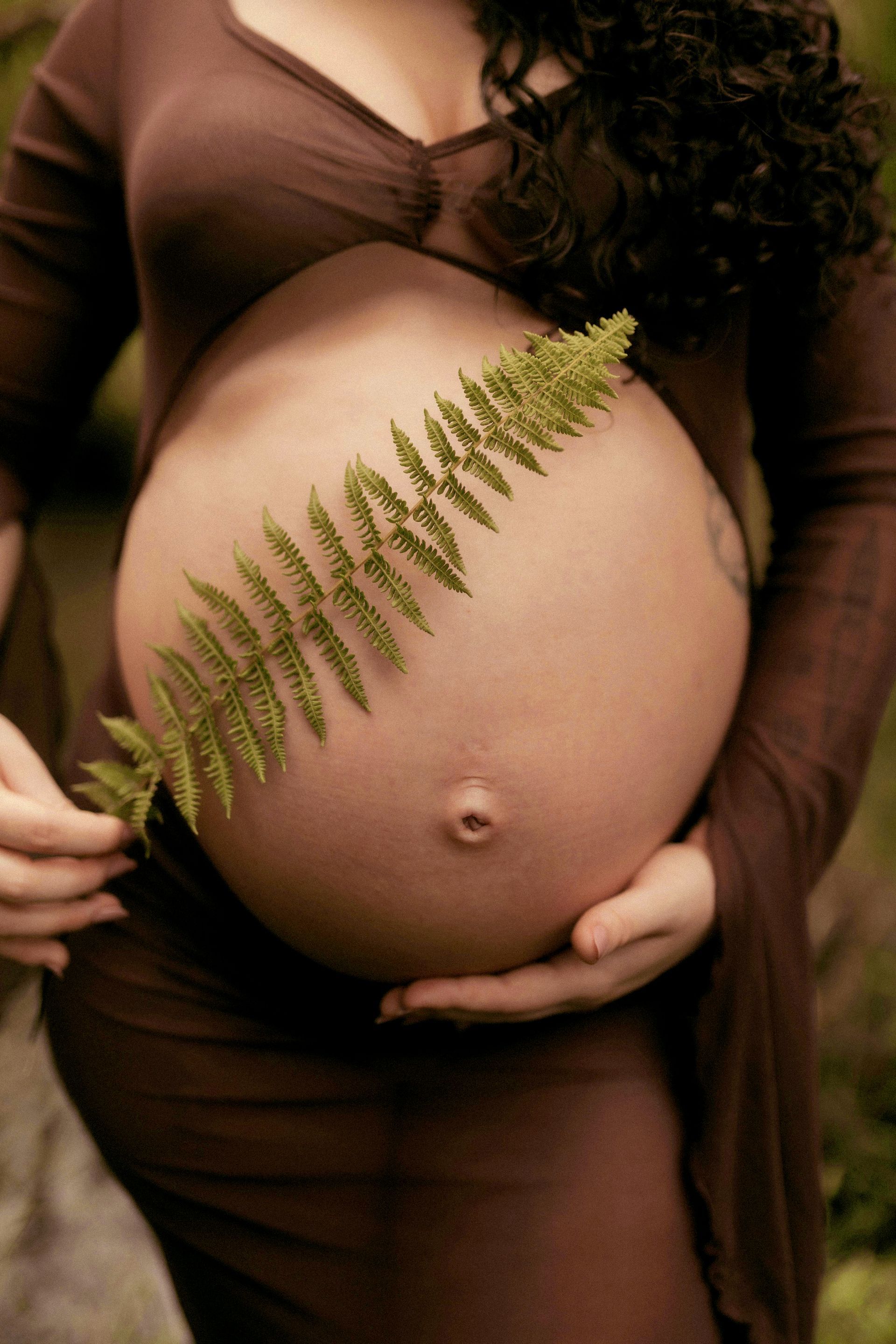 Pregnant person in brown dress holding a fern across their bare belly.
