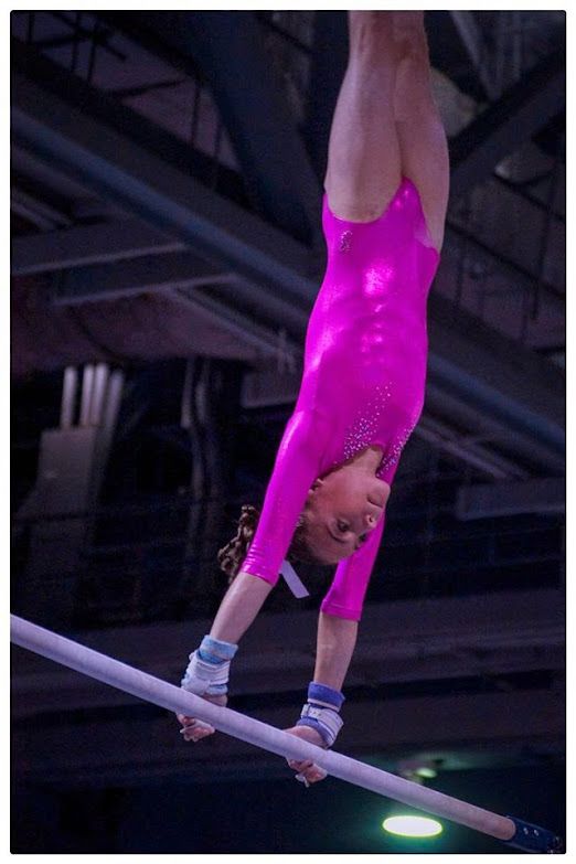Gymnast in a bright pink leotard balances upside down on a beam in an indoor arena.