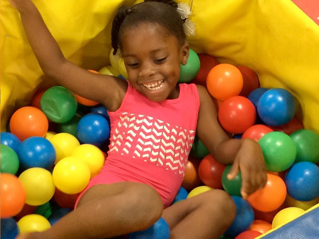 Child smiling in a ball pit with colorful plastic balls and raised arms