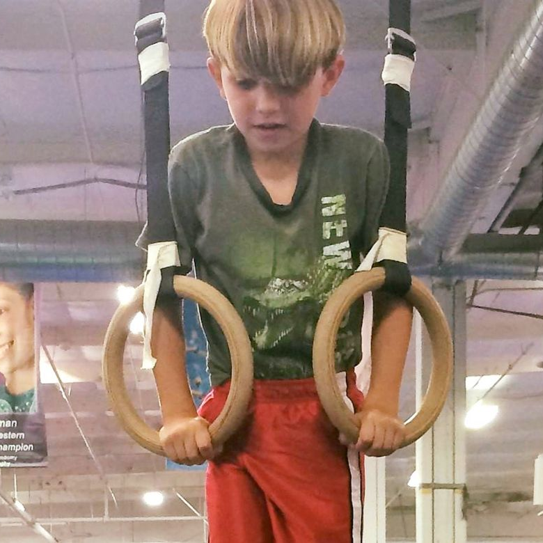 Child doing ring exercises in a gym, gripping wooden rings with a focused expression.