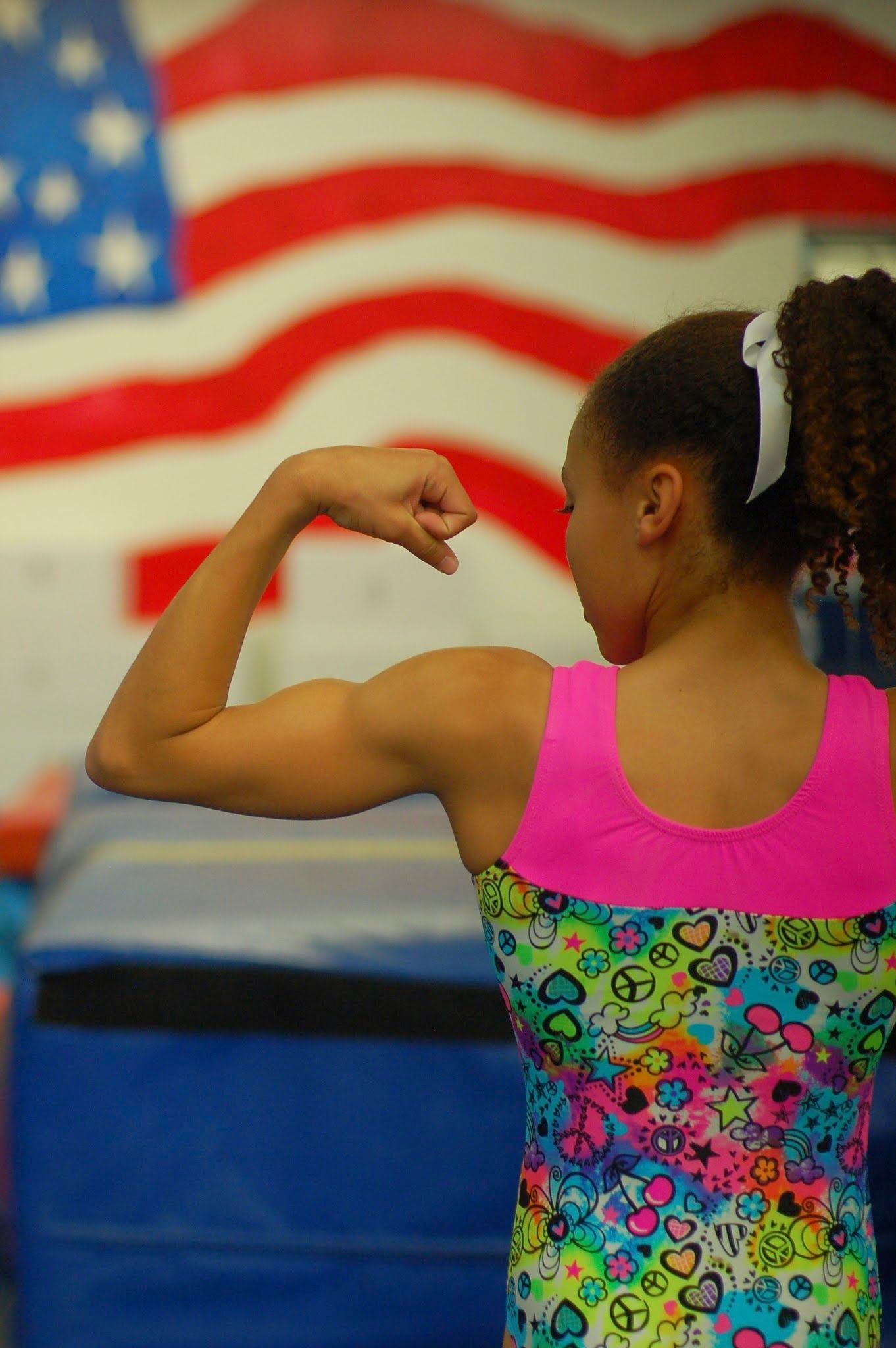 Young gymnast flexing arm in a colorful leotard, with a blurred American flag in the background
