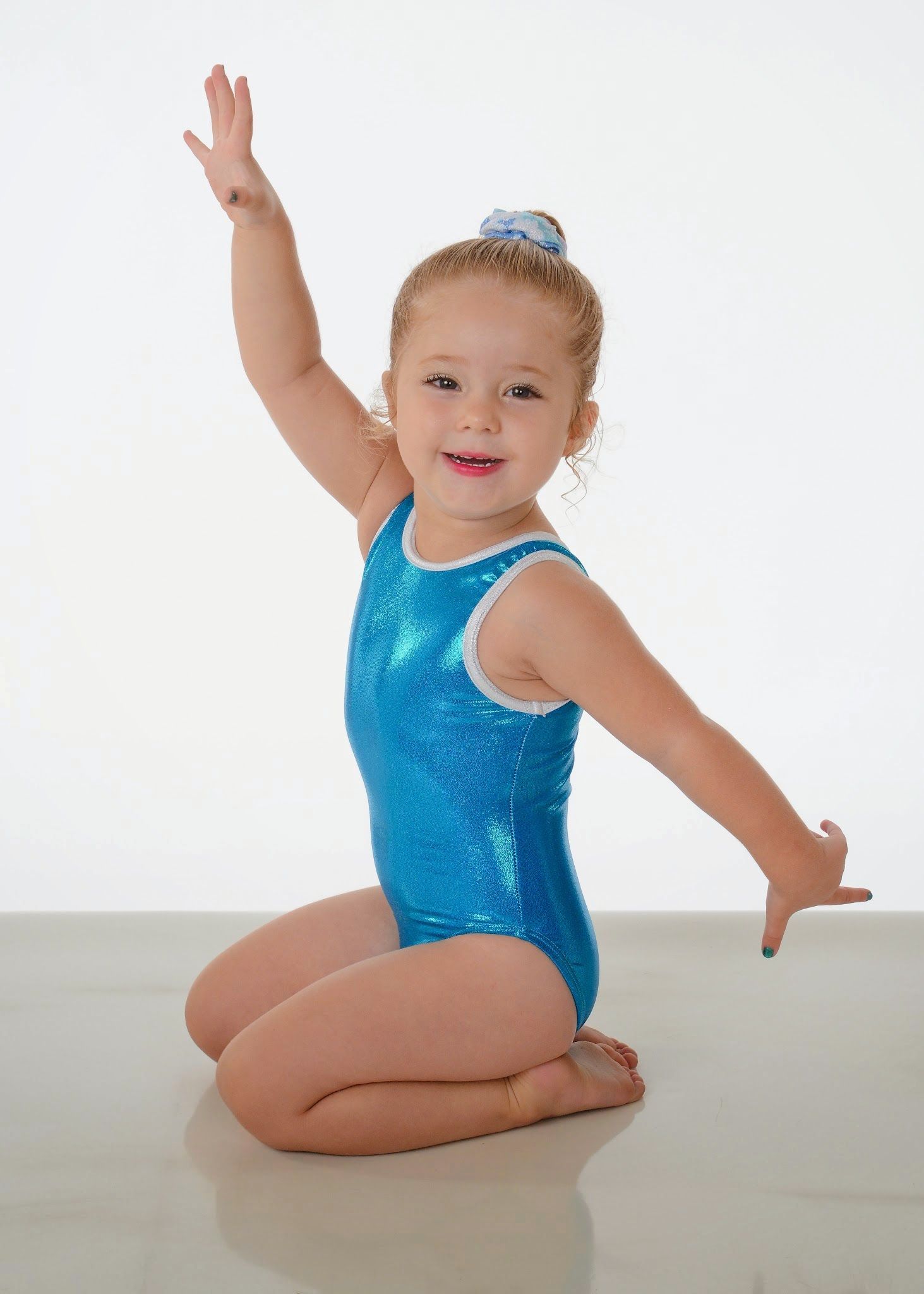 Child in a blue leotard kneeling with arms raised in a white studio, smiling.
