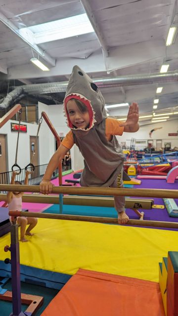 Child in a shark costume climbing on colorful gymnastics equipment in an indoor play gym.