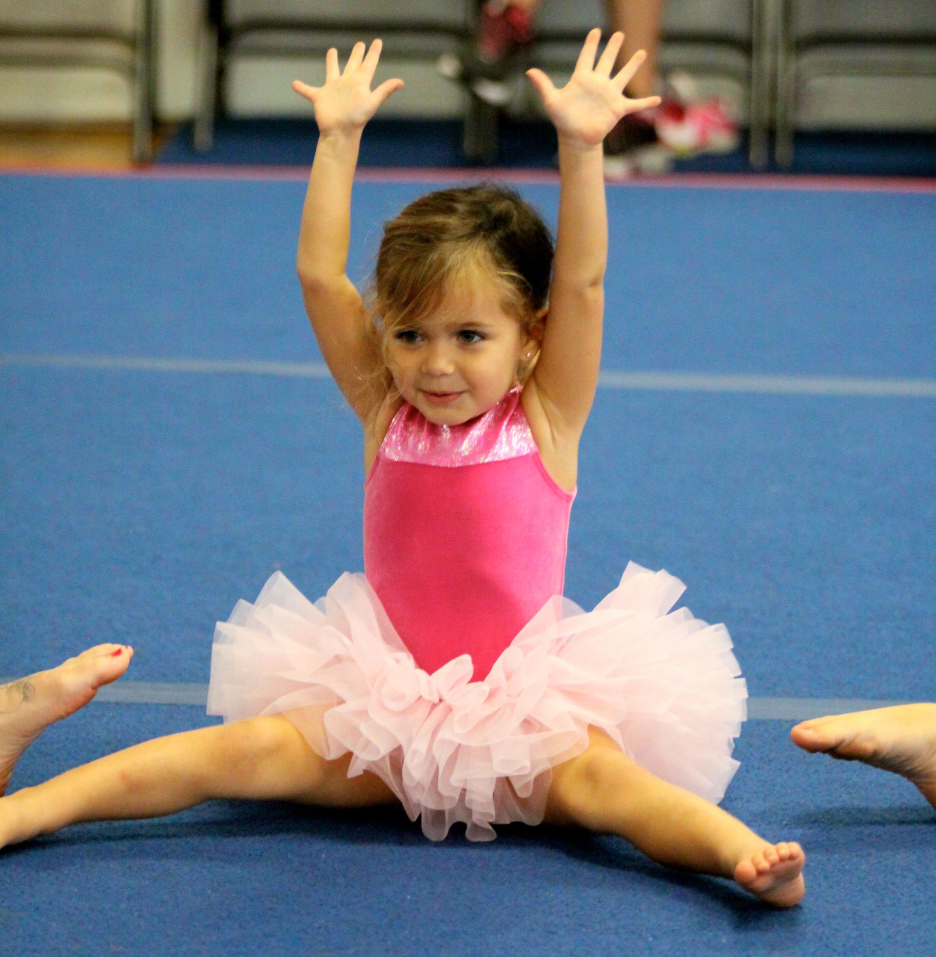 Young child in a pink leotard and tutu doing a split on a gym mat with arms raised.