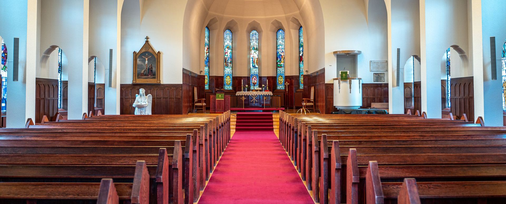 The inside of a church with rows of wooden benches and a red carpet.