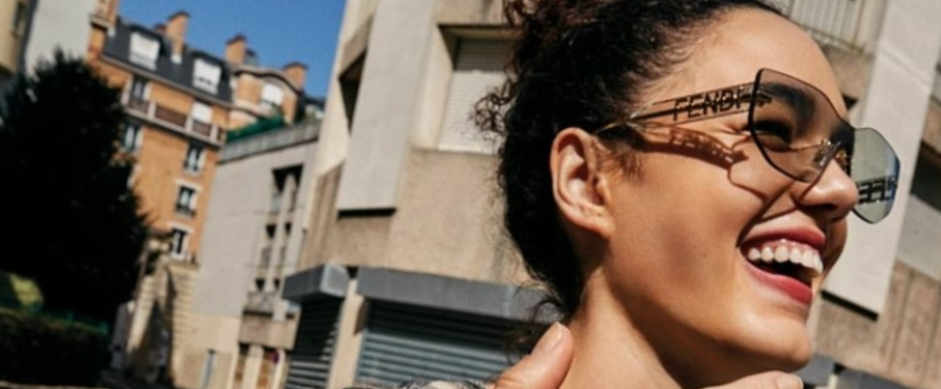 A woman wearing sunglasses is smiling in front of a building.
