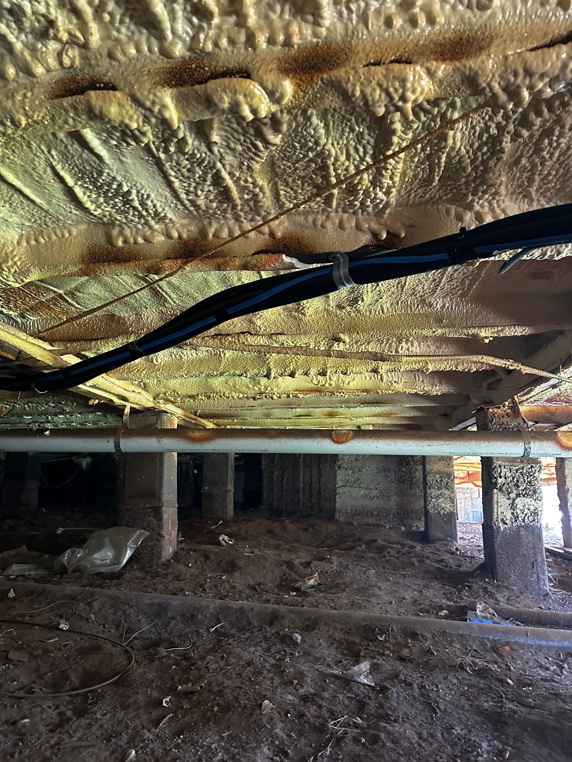 Underside of a house foundation with spray foam insulation, pipes, wiring, and dirt floor — Proshield Insulation & Waterproofing In Shell Cove, NSW