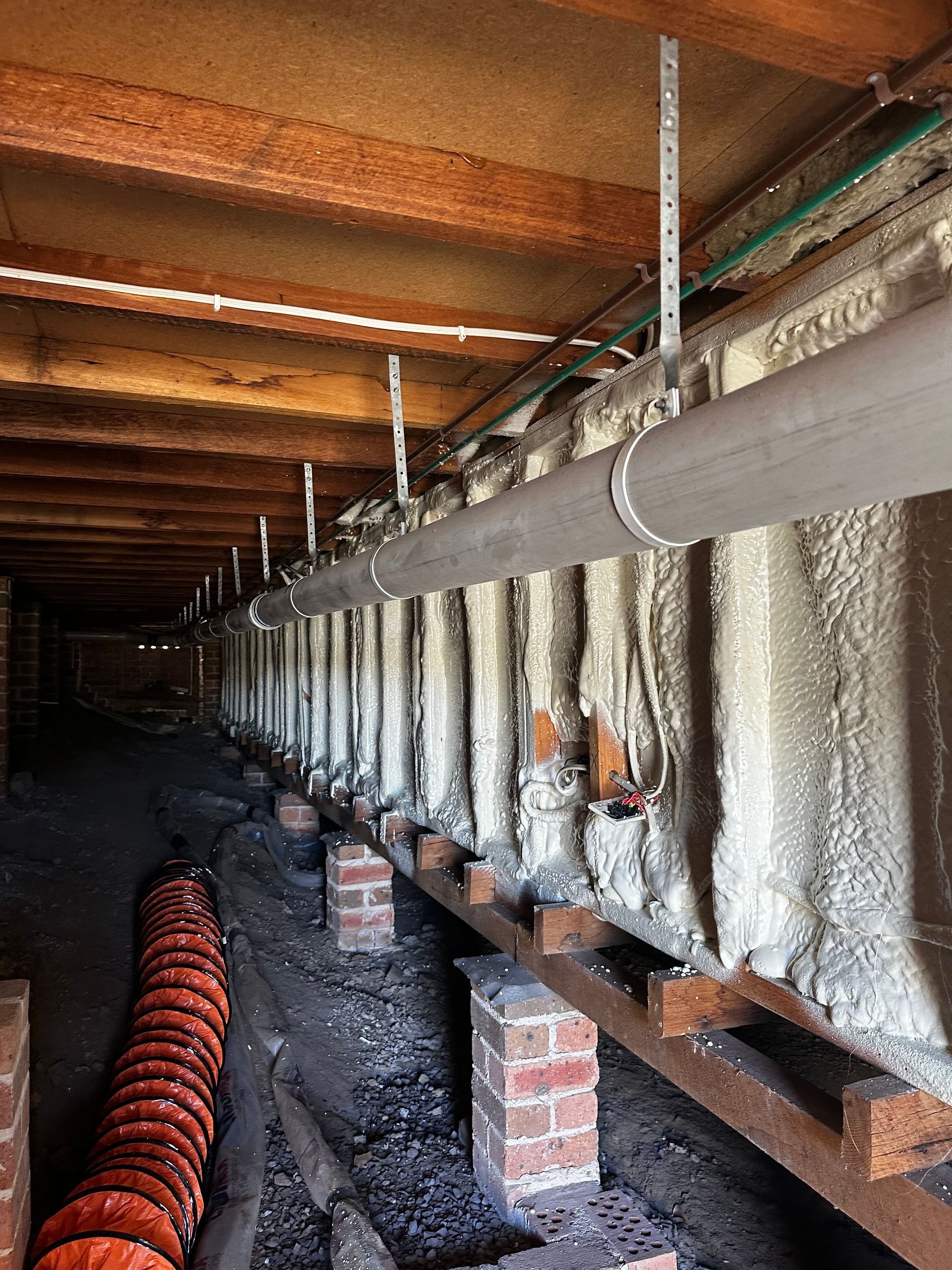 Underneath a house, a long grey pipe is supported by vertical metal rods. Insulation covers the side walls — Proshield Insulation & Waterproofing In Shell Cove, NSW