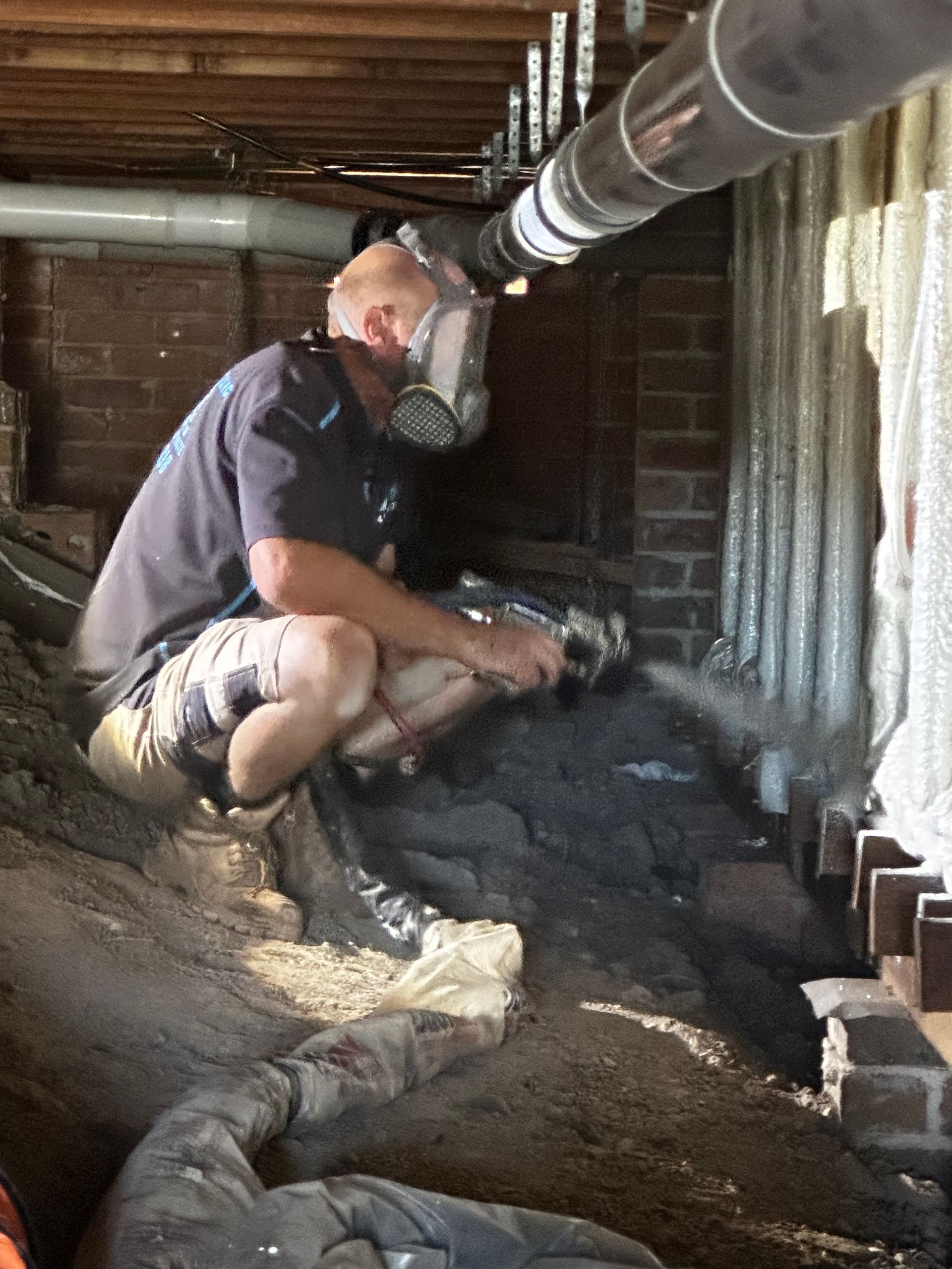 Man in respirator kneels in crawl space, working with gray material; ventilation tube overhead — Proshield Insulation & Waterproofing In Shell Cove, NSW