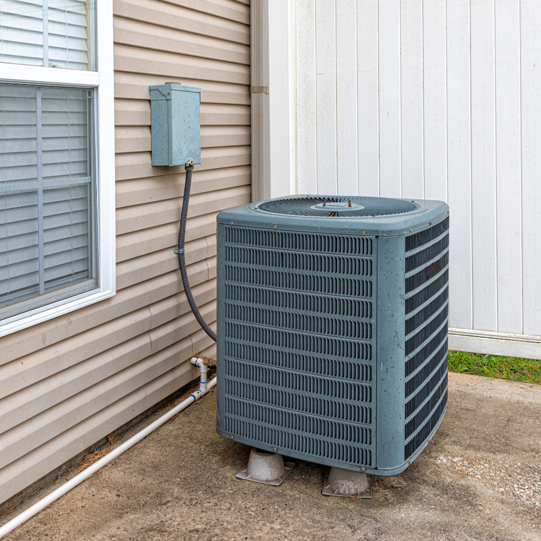 A gray air conditioning condenser unit sitting on a concrete pad next to the tan vinyl siding of a house.