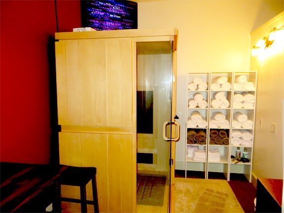 Light-wood infrared sauna cabin with a glass door; shelves of folded towels stand beside it.