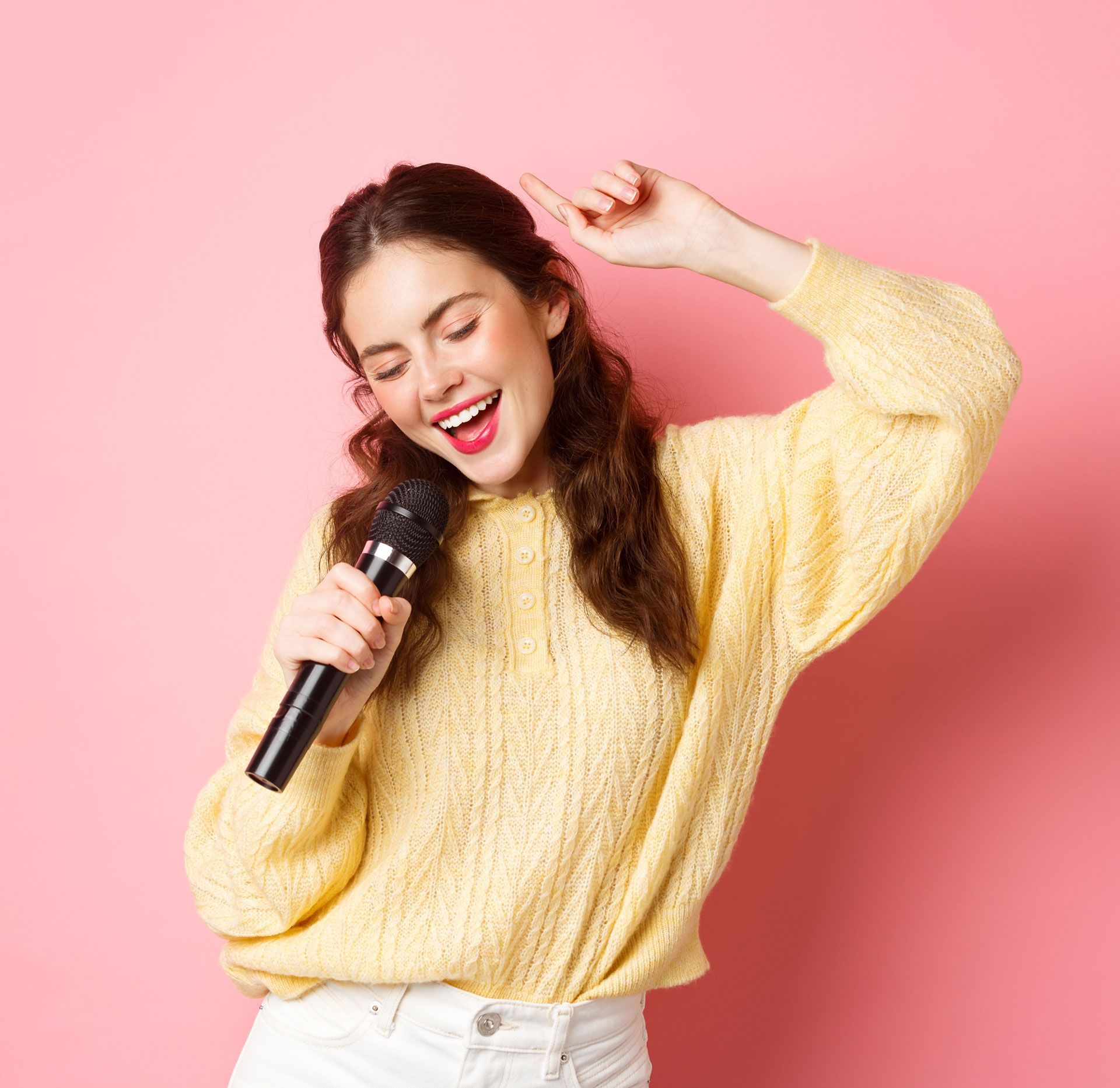 Woman singing into a microphone, wearing a yellow sweater and white pants, smiling against a pink backdrop.