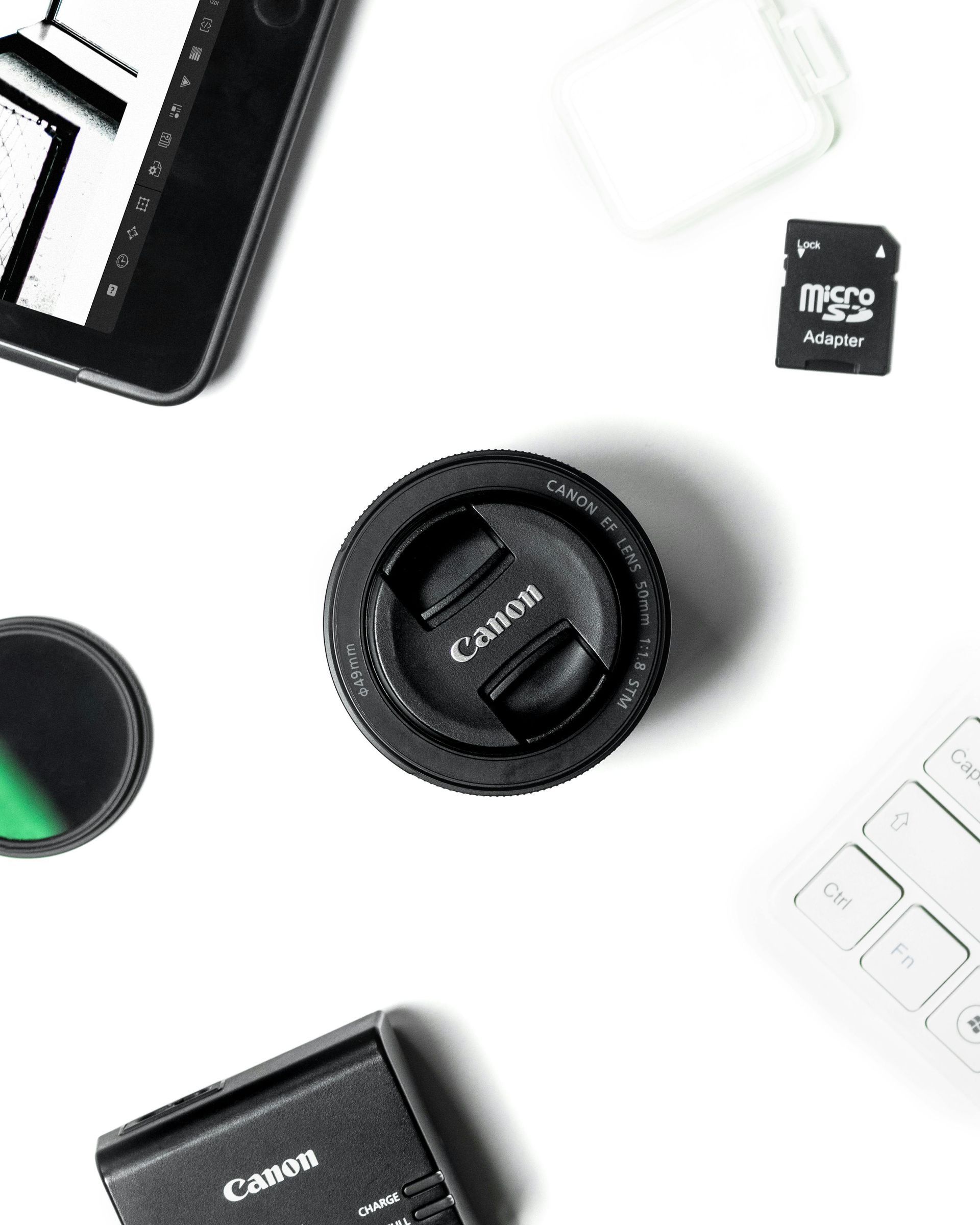 Overhead shot of camera equipment: Canon lens, tablet, keyboard, SD card, battery charger, and filter on white.