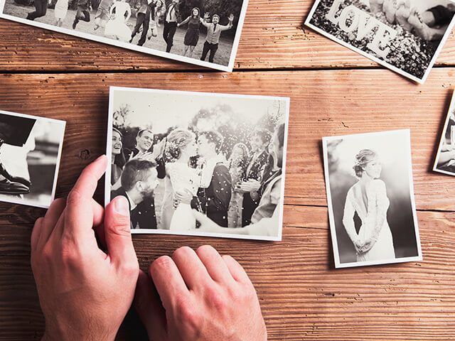 Hands holding a printed photo of a couple kissing amidst a crowd; other photos on wood table.